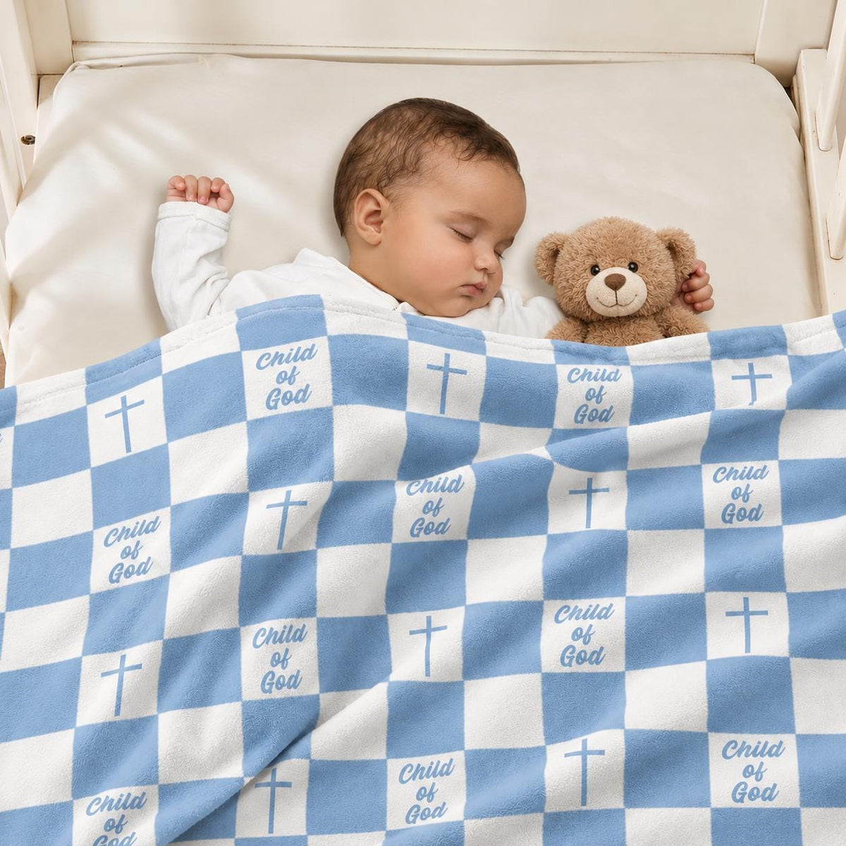 Blue and white Christian baby blanket with Child of God design draped over a sleeping baby in a cozy nursery, featuring a blue gingham pattern with crosses and Child of God lettering, shown with a soft brown teddy bear. ELLO HOME BABY