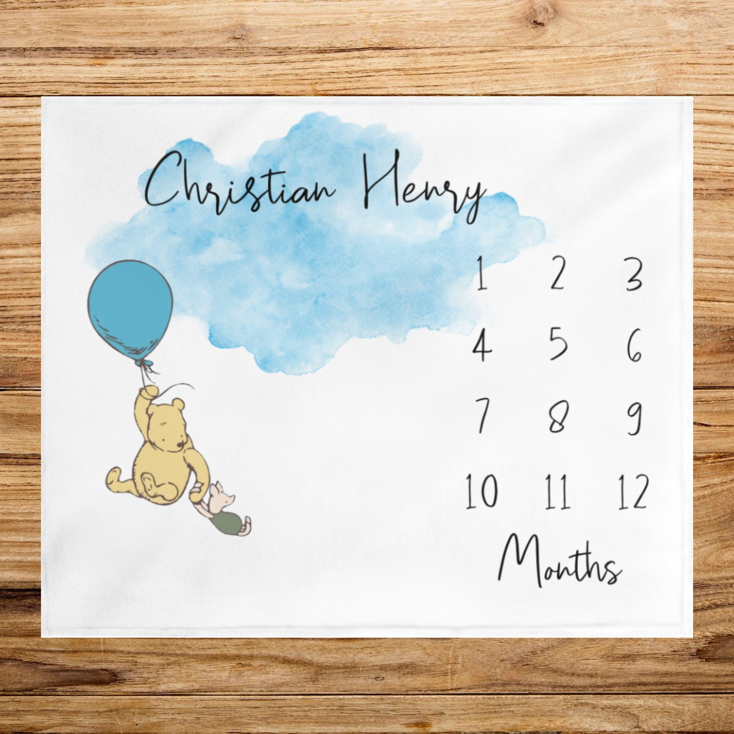 Storybook bear baby milestone blanket with a blue watercolor cloud and a blue balloon. Winnie Classic Bear. The blanket has a chart with numbers 1 through 12 for tracking a baby's monthly growth. The text "Christian Henry" is at the top of the blanket.