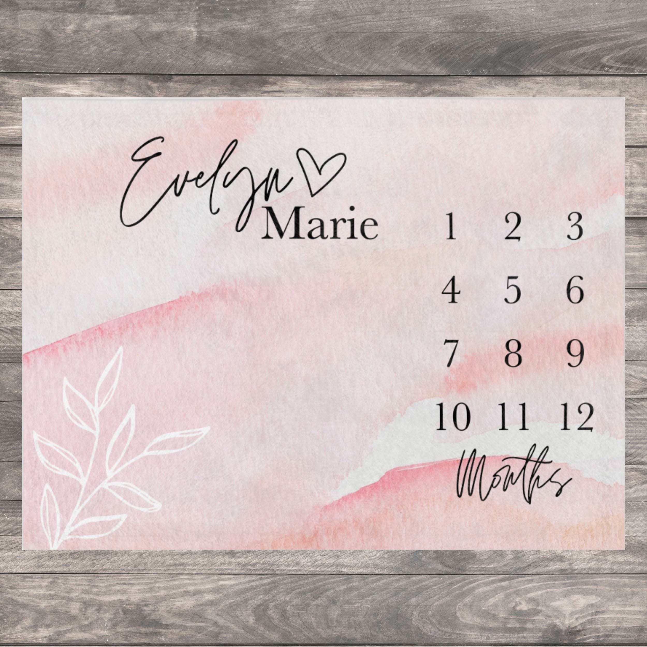 Pink watercolor background with a white floral design and a black handwritten name with a heart symbol.  The image also includes a black number chart for 12 months. Celebrate every precious milestone with a beautifully personalized baby milestone blanket. perfect for capturing your baby’s first year memories in photos. An elegant watercolor milestone blanket features a soft blush pink design with modern typography and a custom baby name.