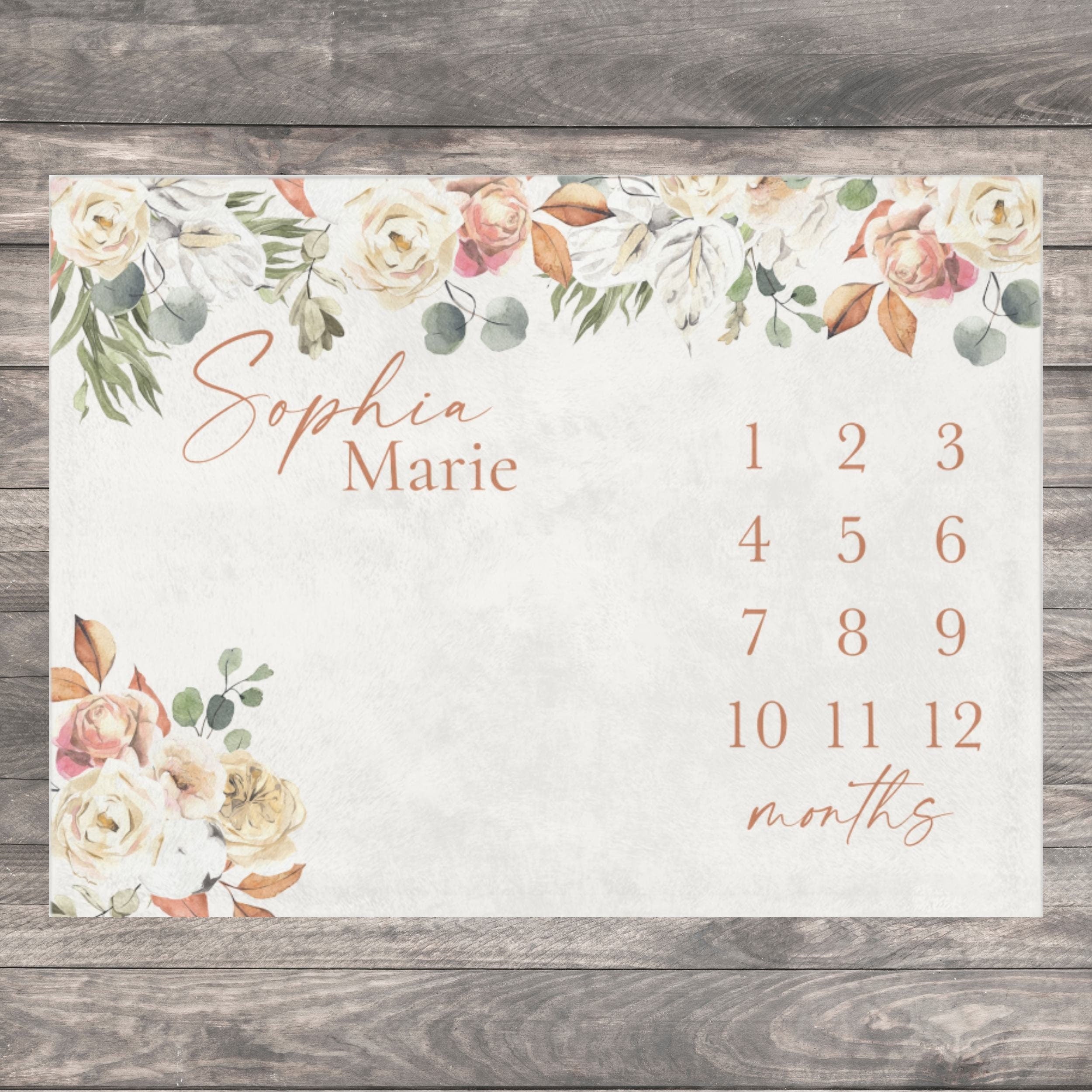 A rectangular milestone blanket featuring a watercolor floral arrangement in cream, peach, and sage green tones. The text reads Sophia Marie, followed by the numbers 1 through 12 and the word months in a copper-colored serif font.