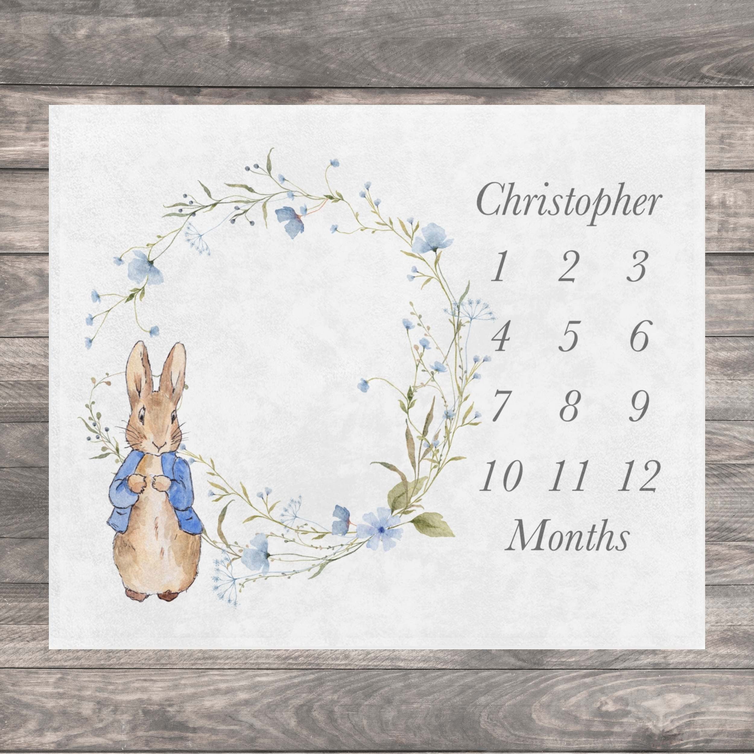 Vintage Rabbit Baby Milestone blanket featuring a watercolor illustration of a blue floral wreath with a Peter Rabbit character in the center. The baby growth chart includes the name "Christopher" and numbers 1 through 12 to track a baby's growth in months.