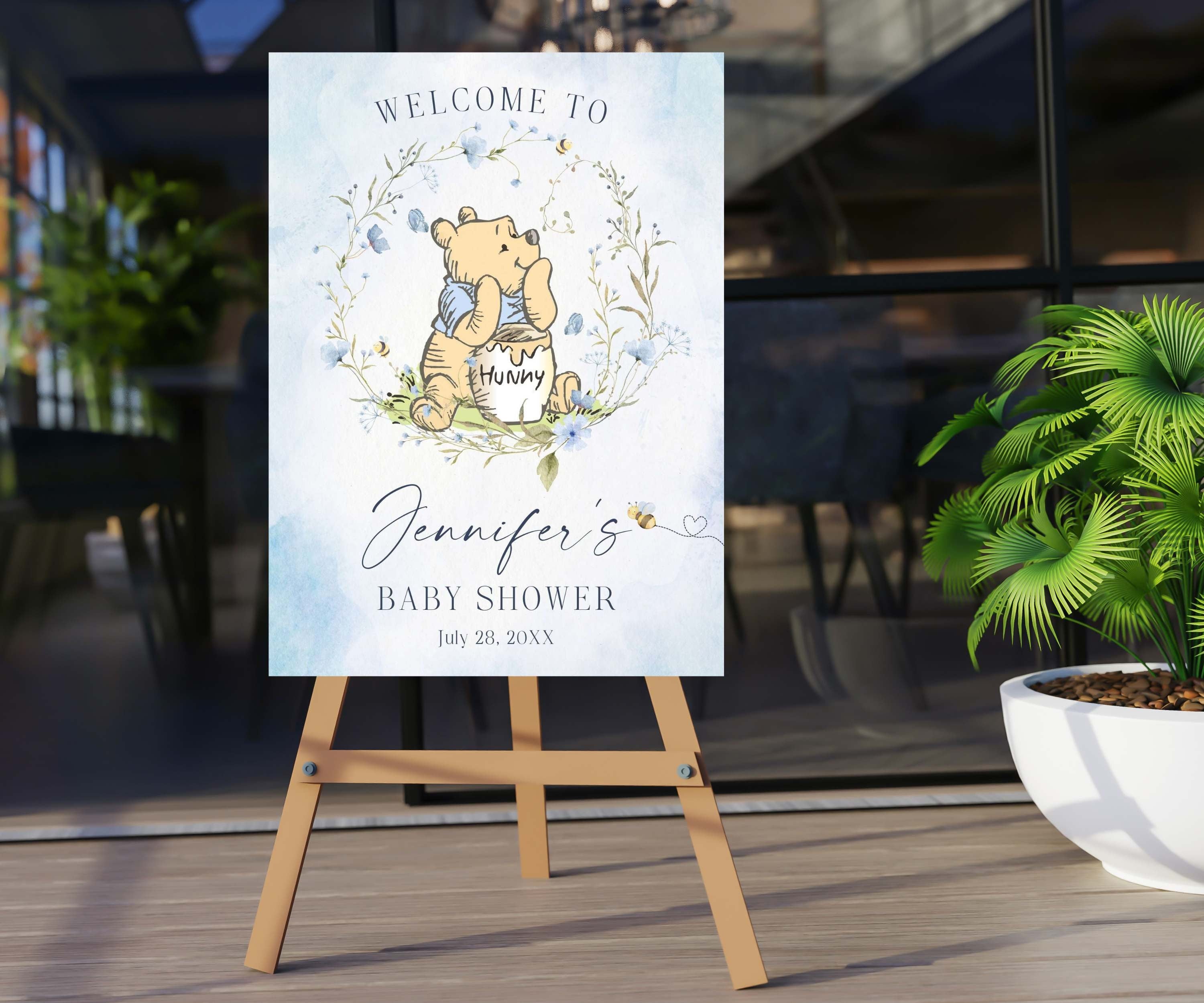 May include: Welcome to Jennifer's Baby Shower sign with a Winnie Bear with a blue shirt illustration. The sign features a blue watercolor background with a wreath of blue flowers and green leaves. Pooh bear is holding a jar of honey and there are bees flying around him. The text on the sign reads "Welcome to Jennifer's Baby Shower July 28, 20XX".