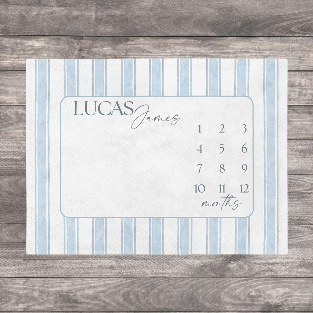 Blue Heirloom Stripe Baby Milestone Blanket. Baby Milestone Blanket Boy, Personalized Month Blanket Blue Striped, Preppy Monthly Growth Mat, Custom Baby Shower Gift. A rectangular milestone blanket with a light blue and white striped border. The center features a light blue frame with the name "LUCAS James" and numbers 1-12 with the word "months" below.