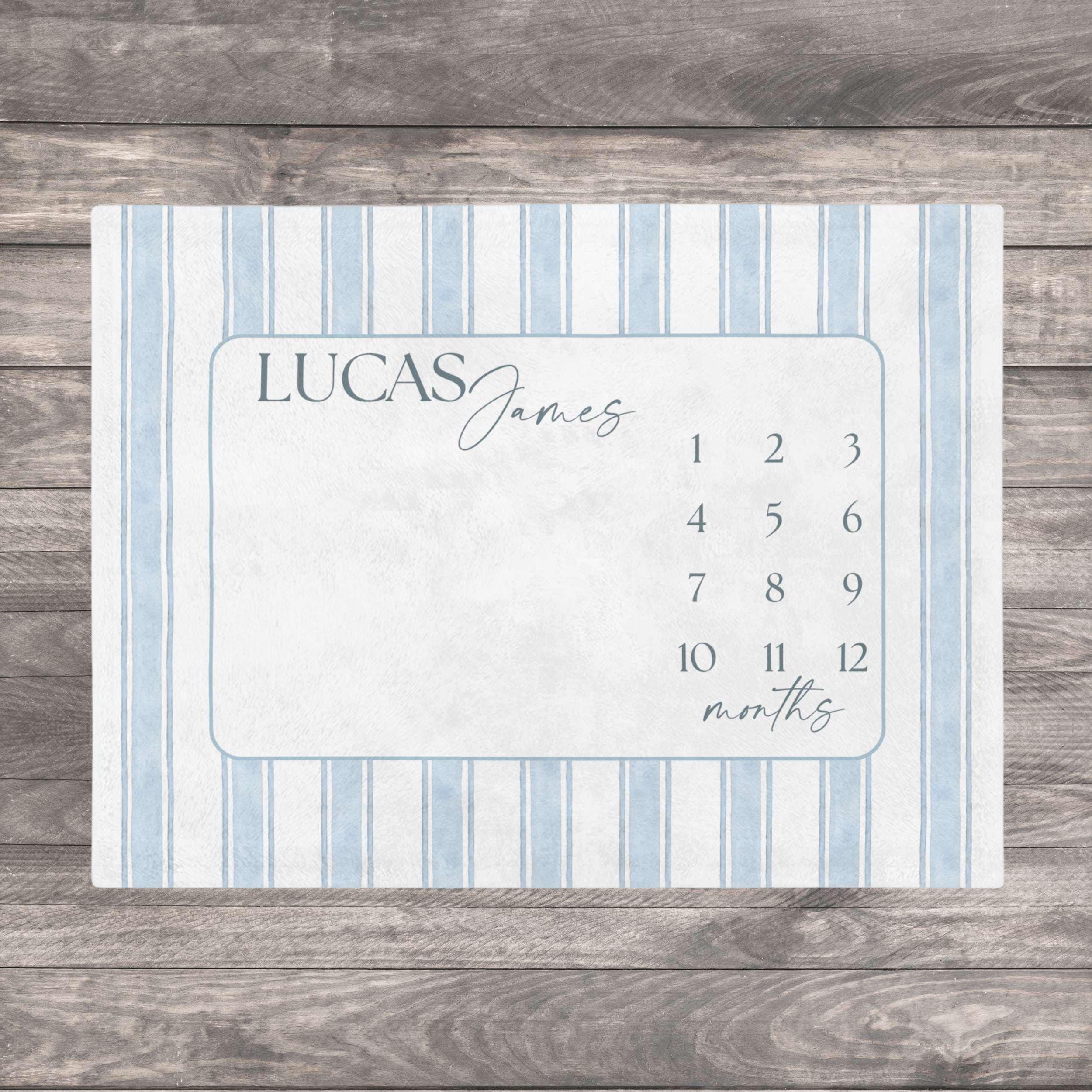 Blue Heirloom Stripe Baby Milestone Blanket. Baby Milestone Blanket Boy, Personalized Month Blanket Blue Striped, Preppy Monthly Growth Mat, Custom Baby Shower Gift. A rectangular milestone blanket with a light blue and white striped border. The center features a light blue frame with the name "LUCAS James" and numbers 1-12 with the word "months" below.