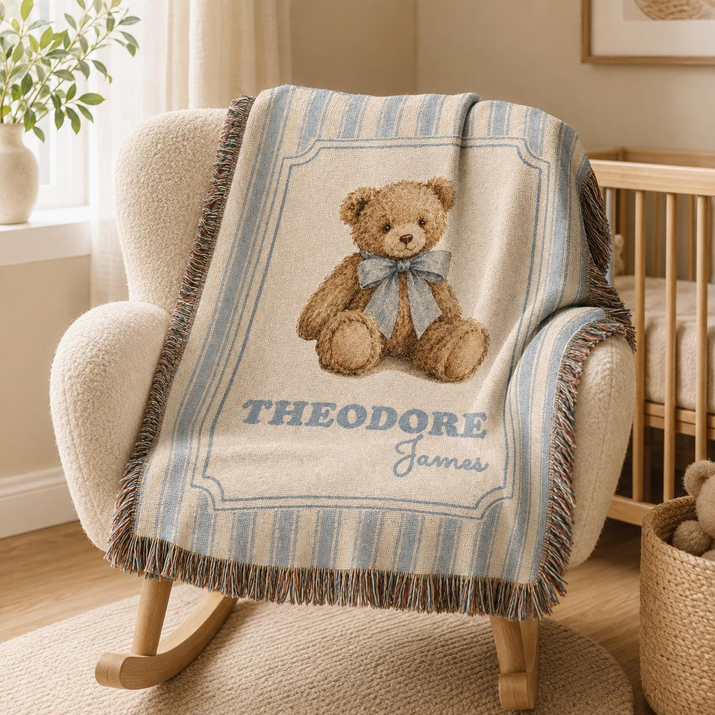 Personalized blue striped cotton woven baby blanket laid on a chair inside a neutral nursery, featuring a brown teddy bear with a dusty-blue bow, custom name, framed design and fringed edges.