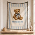 Personalized Custom Name blue striped cotton woven baby blanket hanging in a bright neutral nursery, featuring a medium-brown teddy bear with a dusty-blue bow, framed design and fringed edges.