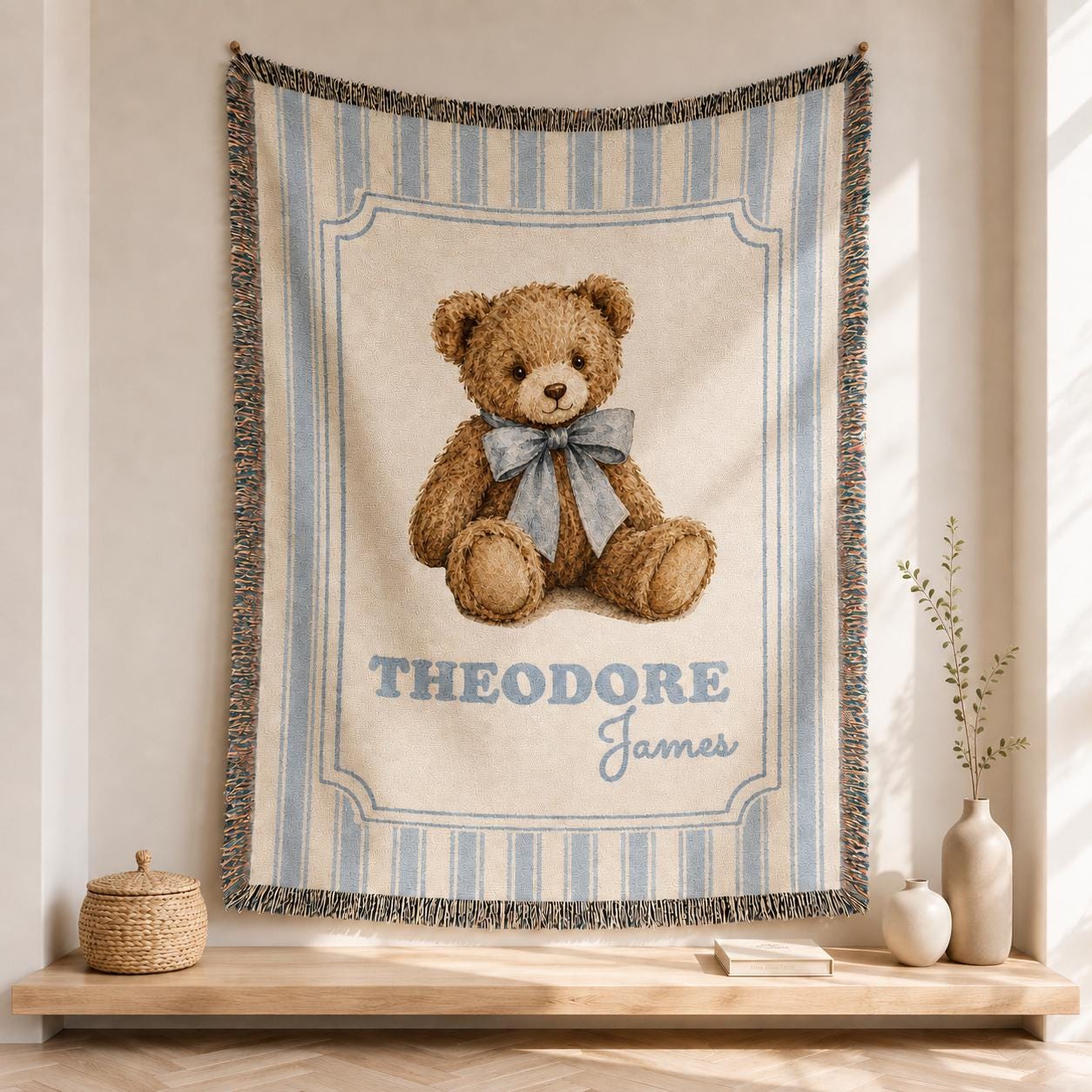 Personalized Custom Name blue striped cotton woven baby blanket hanging in a bright neutral nursery, featuring a medium-brown teddy bear with a dusty-blue bow, framed design and fringed edges.