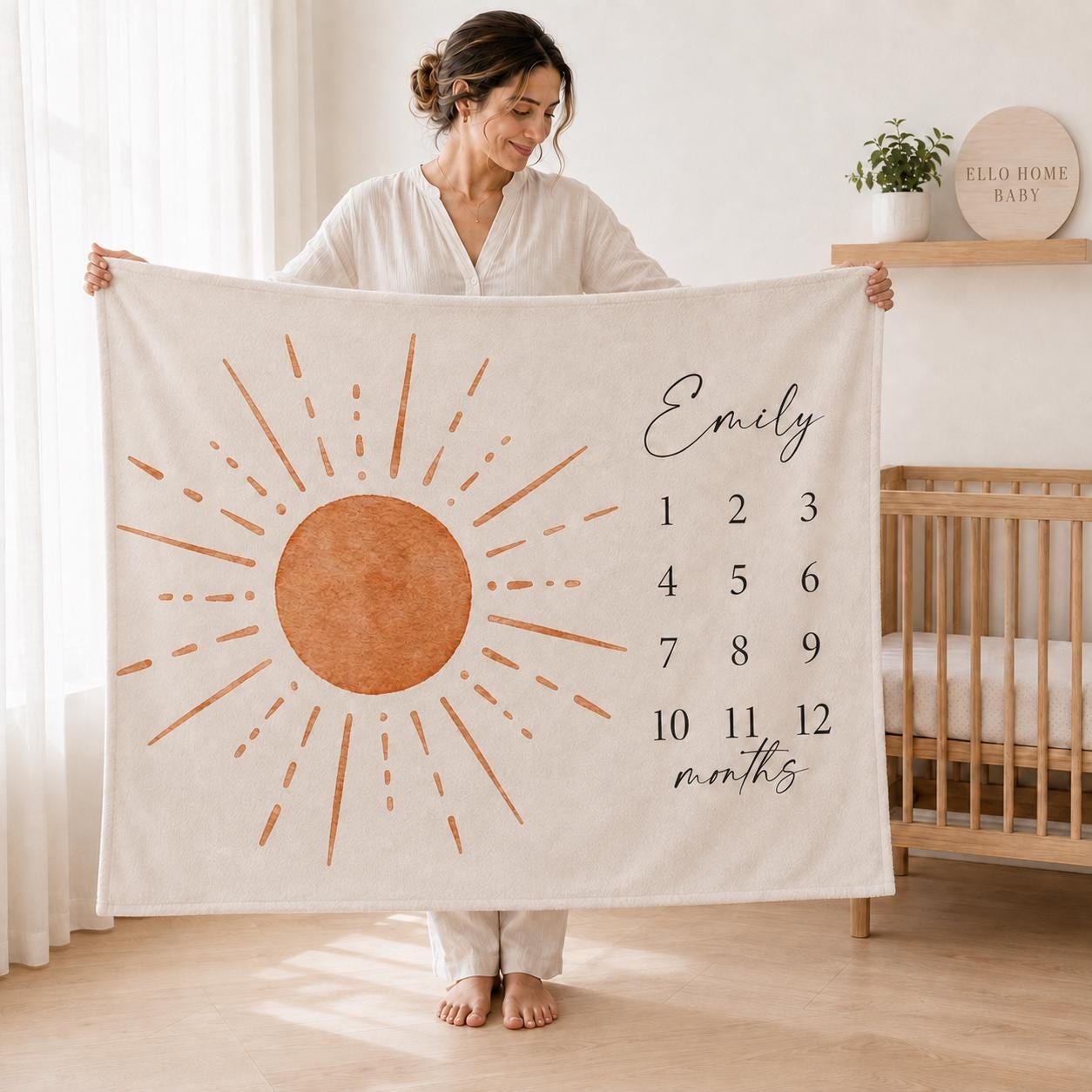 Mother holding a 60 by 50 inch personalized sun baby milestone blanket in a bright nursery with a light wood crib.