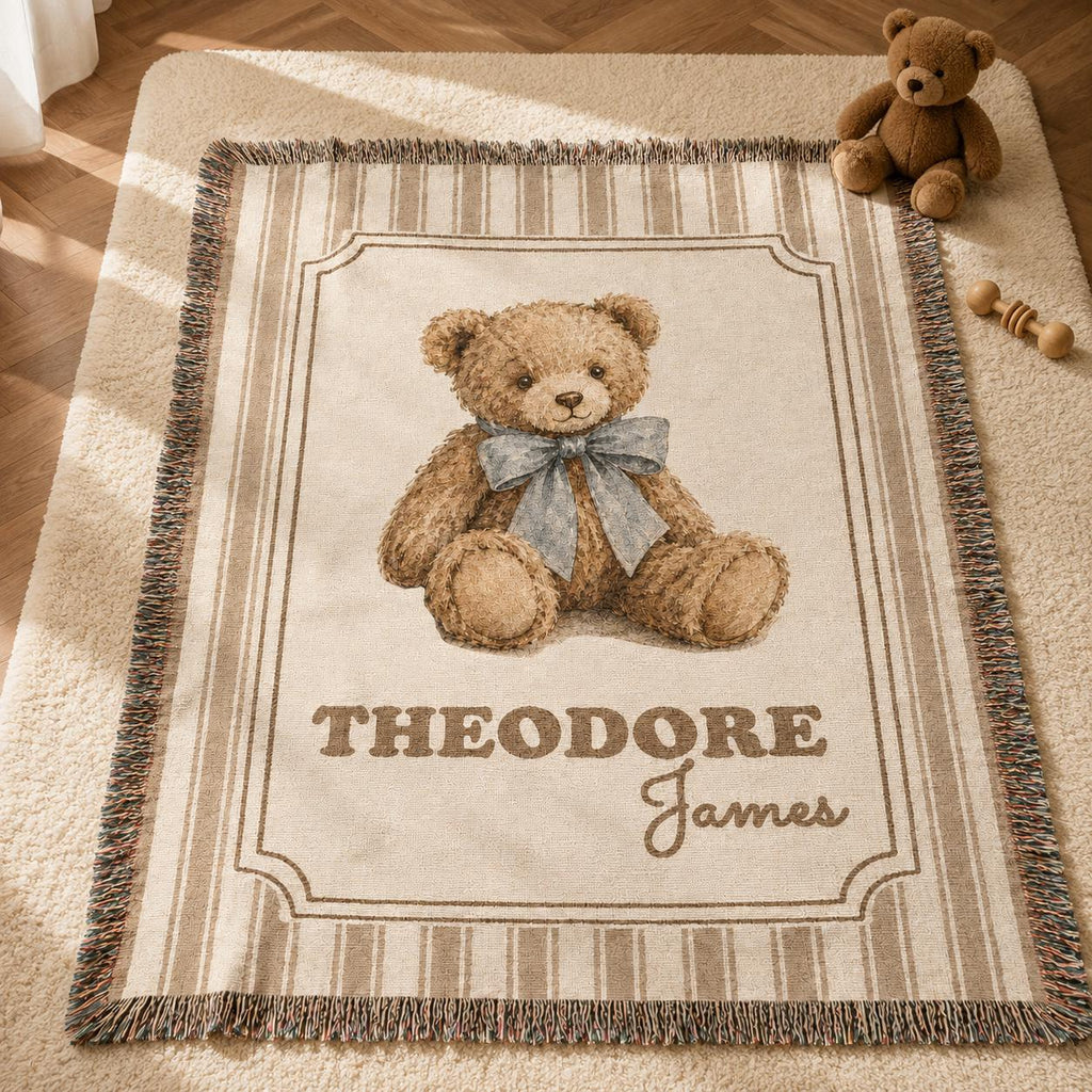 A personalized vintage-style woven nursery blanket featuring a realistic illustration of a brown teddy bear wearing a dusty blue bow. The name 'Theodore James' is customized at the bottom. The blanket is 100% cotton, has vertical brown and beige stripes and a fringed edge, displayed on a wooden herringbone floor next to a plush teddy bear and a potted plant with natural sunlight, a plush bear nursery theme.
