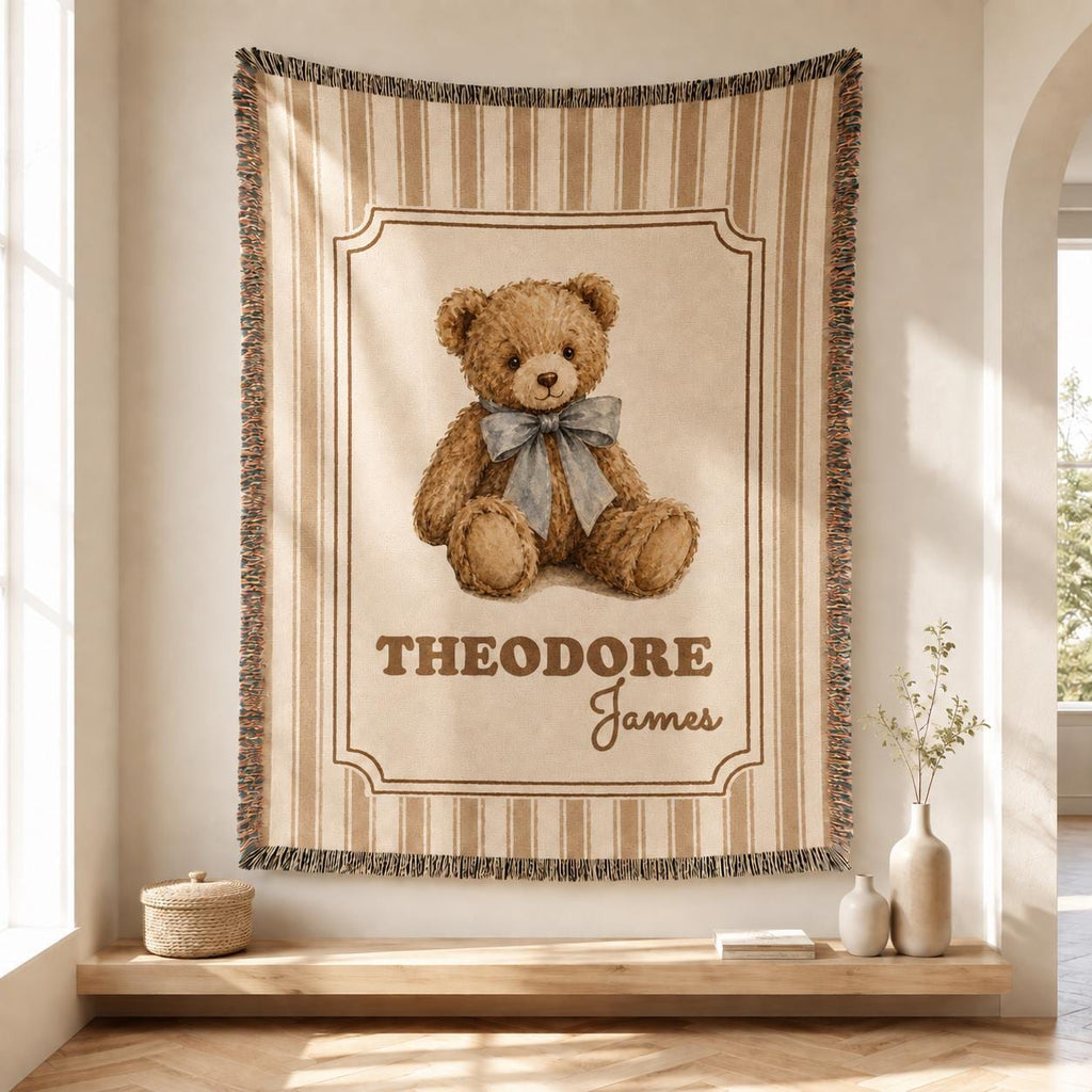 Personalized brown striped cotton woven baby blanket hanging in a bright neutral nursery, featuring a medium-brown teddy bear with a dusty-blue bow, Theodore James name, framed design and fringed edges.