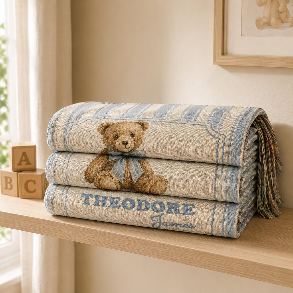 Personalized blue striped cotton woven baby blanket folded nicely on a shelf display, Stacked on a floating nursery shelf next to ABC wooden blocks, in a bright neutral nursery, featuring a classic brown teddy bear with a dusty-blue bow, featuring a custom baby name. 100% cotton woven baby blanket.