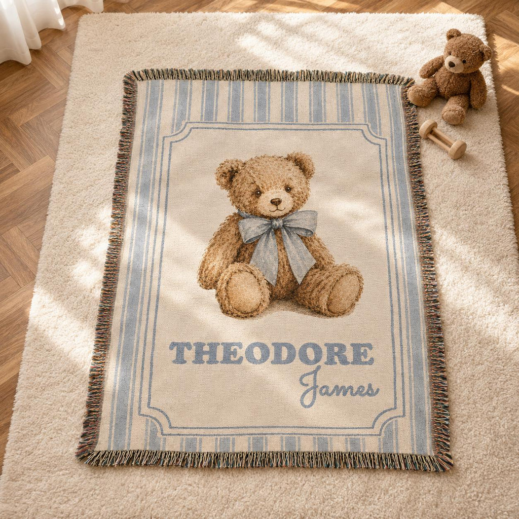 A personalized vintage-style woven nursery blanket featuring a realistic illustration of a brown teddy bear wearing a dusty blue bow. The name 'Theodore James' is customized at the bottom. The blanket is 100% cotton, has vertical blue and beige stripes and a fringed edge, displayed on a wooden floor next to a plush teddy bear with natural sunlight. Classic old money teddy bear nursery theme.