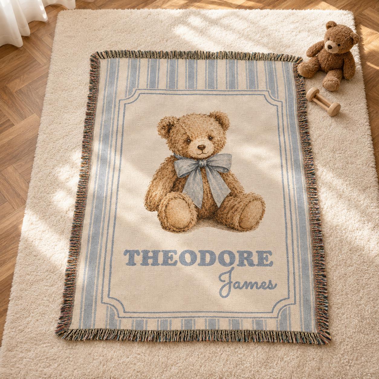 A personalized vintage-style woven nursery blanket featuring a realistic illustration of a brown teddy bear wearing a dusty blue bow. The name 'Theodore James' is customized at the bottom. The blanket is 100% cotton, has vertical blue and beige stripes and a fringed edge, displayed on a wooden floor next to a plush teddy bear with natural sunlight. Classic old money teddy bear nursery theme.