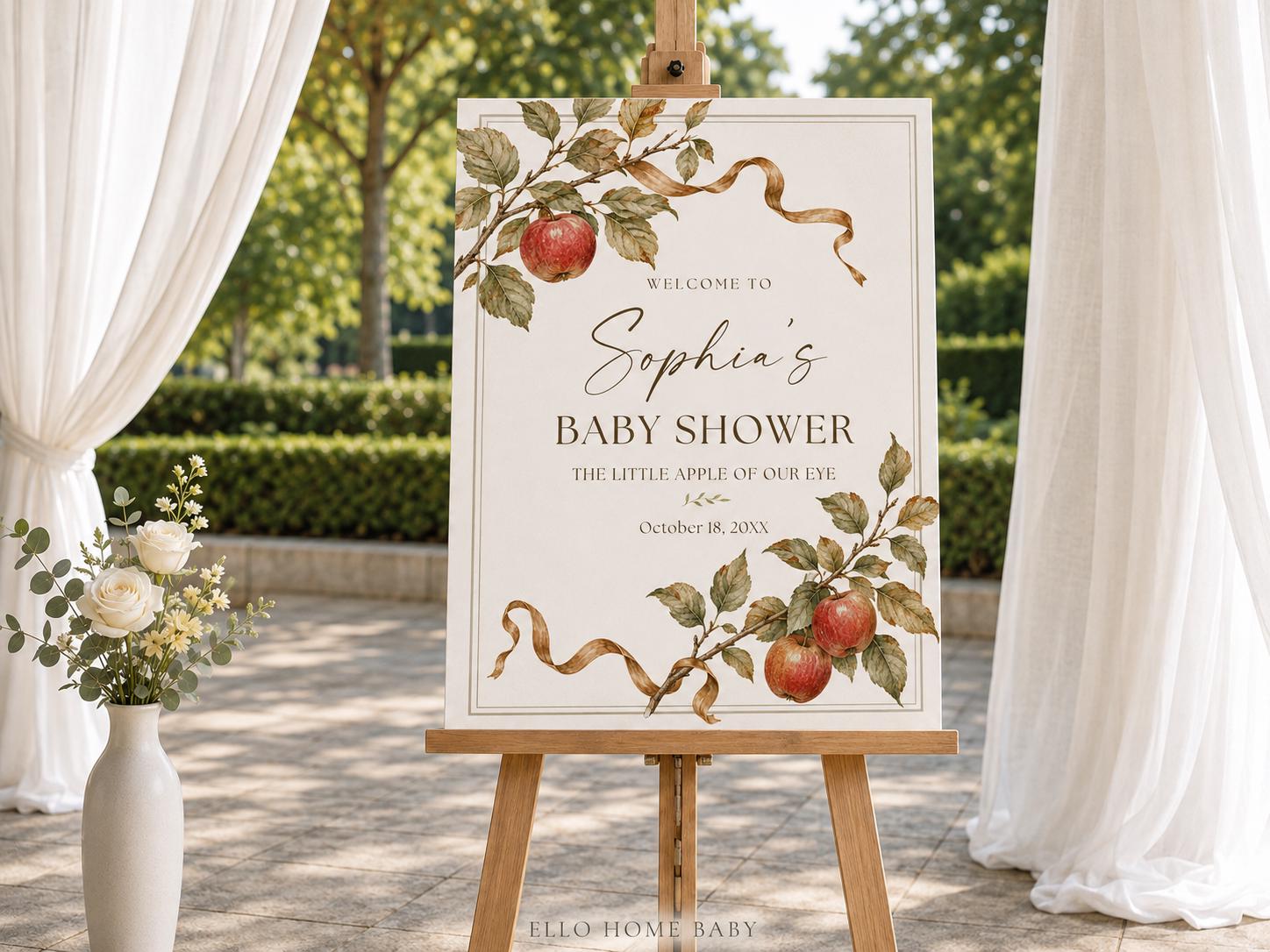 Fall apple baby shower foam-board sign displayed on a wooden tripod easel at an outdoor garden celebration.