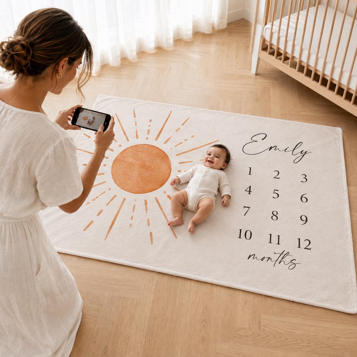 Mother photographing her smiling baby on a plush minky milestone blanket with a personalized name, sun design and month numbers.