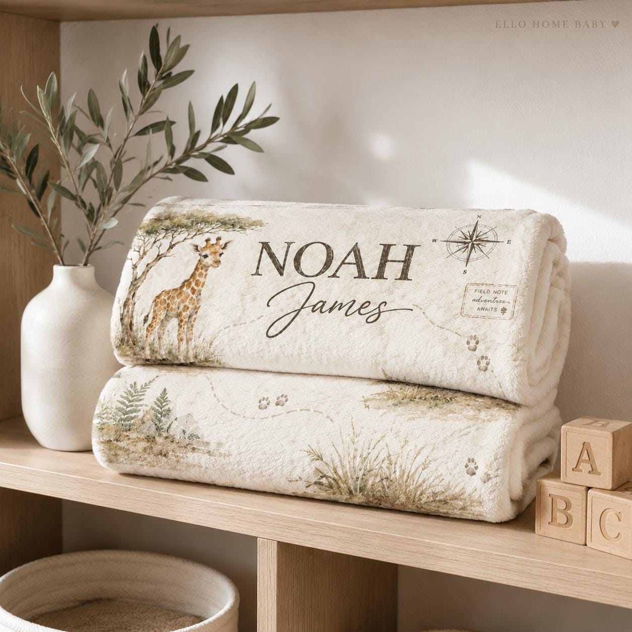 Folded personalized safari milestone blanket on a nursery shelf showing a watercolor giraffe, compass, paw prints and custom baby name.