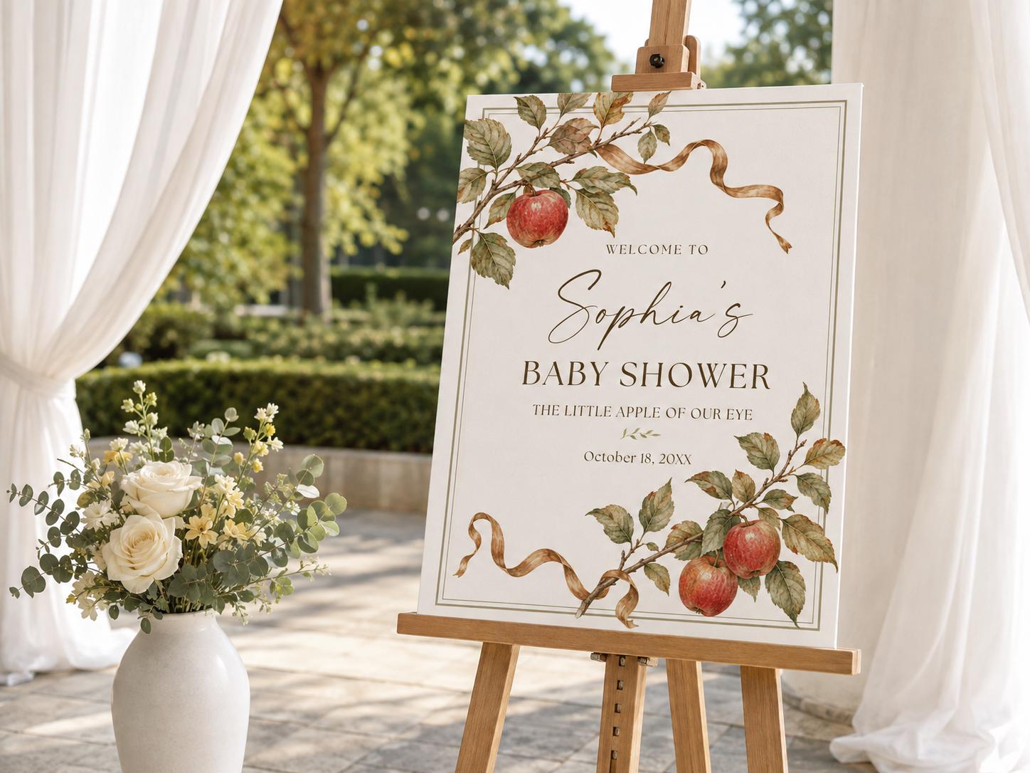 Personalized Little Apple of Our Eye baby shower welcome sign with watercolor red apples, autumn leaves and flowing brown ribbons displayed on a wooden easel.